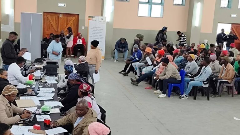 Applicants at the SASSA office completing grant applications, some seated, others submitting documents to SASSA representatives at service desks, SASSA July 2025 Grant Application Apply Now